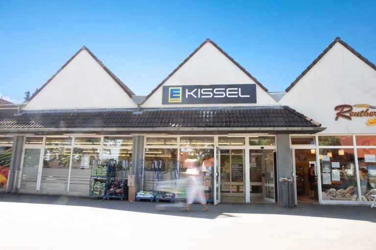 Zu sehen ist eine Fotografie des Außengeländes des EDEKA Kissel Marktes in Klingenmünster. Über dem Markteingang hängt das EDEKA Kissel-Logo. Vor dem Gebäude stehen Rollcontainer mit Blumenerde. Rechts neben dem Markt ist eine Reuther Bäckerei zu sehen. Vom Markt wegbewegend zu sehen, ist ein Kunde mit Einkaufswagen in Bewegung.