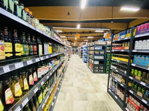 Zu sehen ist eine Fotografie des Innenbereiches des Edeka Kissel Marktes in Edesheim. Das Foto zeigt gefüllte Regale. Links im Bild stehen verschiedene Öle und Essige. In den Regalen rechts im Bild stehen Energydrinks und Bier. Am Ende des Ganges zu erkennen sind Kunden mit Einkaufswagen in Bewegung.