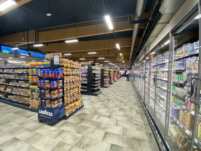 Zu sehen ist eine Fotografie eines Edeka Kissel Marktes, bei der schräg in den Markt fotografiert wurde. Rechts im Bild abgebildet ist eine Kühltheke gefüllt mit frischer Milch, Joghurt, die sich über die ganze Wandseite zieht. Links sind Regale mit Kaffee, Toast, Aufbackbrötchen zu erkennen. Im Hintergrund lassen sich Regale mit Müsli, Marmelade und Nudeln erahnen.
