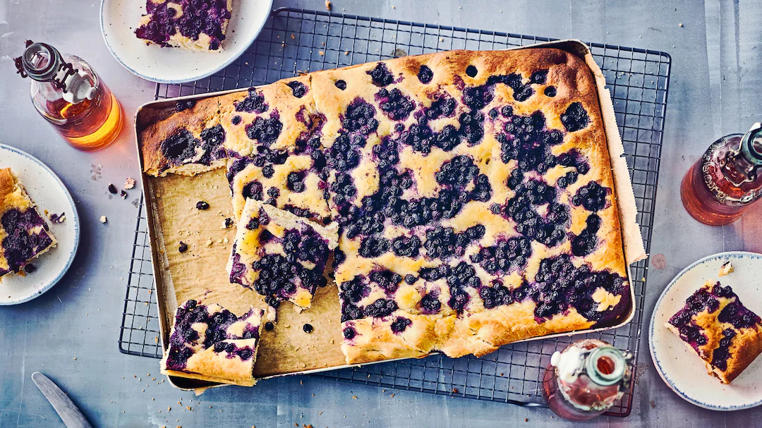 Zu sehen ist ein rechteckiger Blaubeer-Butterkuchen auf einem Backblech, das auf einem schwarzen Gitterrost liegt. Der Kuchen ist goldbraun gebacken und mit vielen frischen Blaubeeren bedeckt. Einige Stücke wurden bereits herausgeschnitten und liegen auf weißen Tellern um das Backblech herum. Es stehen drei Glasflaschen mit einem rötlichen Getränk auf dem Tisch. Der Hintergrund ist eine graue Tischoberfläche mit Kuchenkrümmeln und kleinen Flecken.