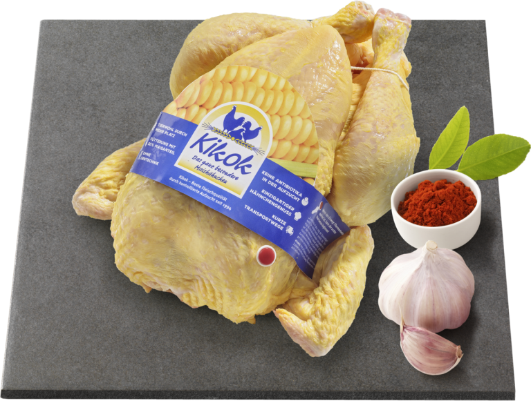Zu sehen ist ein gelbes Kikok Hähnchen platziert auf einer grauen Platte. Am Hähnchen befestigt ist ein blaues Etikett, welches über die Besonderheiten des Kikok Hähnchens informiert. Rechts auf der Platte abgebildet ist eine Knolle Knoblauch und ein Schüsselchen mit Paprikapulver.