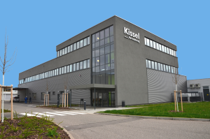 Zu sehen ist die Kissel Verwaltung in der Johannes-Kopp-Straße 12. In großen Buchstaben steht "Kissel Verwaltung" auf der Vorderseite des Gebäudes. Ein Zebrastreifen führt über die Straße zum Gebäudeeingang und zu einer kleinen Grünfläche.